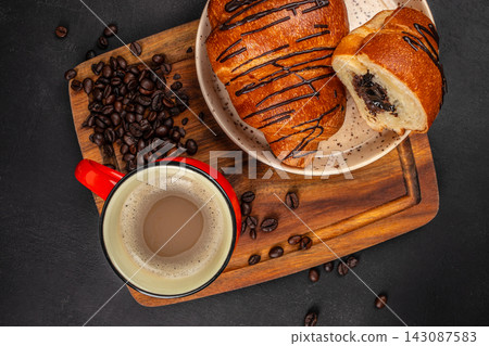 Coffee cup with milk beside a plate of chocolate-filled pastries on a wooden board, scattered coffee beans on a dark textured surface Coffee cup with milk beside a plate of chocolate-filled pastries on a wooden board, scattered coffee beans on a dark textured surface 143087583
