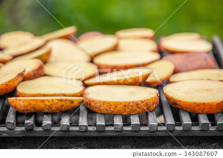 Slices of potatoes grilling on a barbecue grill with a blurred green background, showcasing a summer cooking scene with food preparation outdoors Slices of potatoes grilling on a barbecue grill with a blurred green background, showcasing a summer cooking scene with food preparation outdoors 143087607