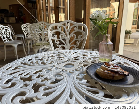 Ornate patio table with pastry and flowers beside sunlit windows in inviting outdoor dining setting Ornate patio table with pastry and flowers beside sunlit windows in inviting outdoor dining setting 143089051