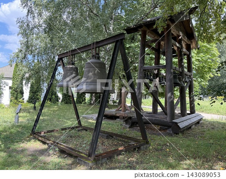 Historic bronze bells hanging from rustic wooden frame in leafy outdoor setting with blue sky and grass Historic bronze bells hanging from rustic wooden frame in leafy outdoor setting with blue sky and grass 143089053