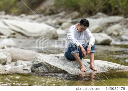 一個神情悲傷的男人坐在河邊的石頭上。 一個神情悲傷的男人坐在河邊的石頭上。 143089476