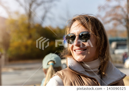 Smiling woman wearing sunglasses enjoys warm autumn sunlight outdoors in candid lifestyle portrait. Golden foliage and soft urban background create relaxed, cheerful seasonal atmosphere Smiling woman wearing sunglasses enjoys warm autumn sunlight outdoors in candid lifestyle portrait. Golden foliage and soft urban background create relaxed, cheerful seasonal atmosphere 143090325
