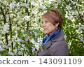 Elegant senior woman with short hair smiling and enjoying spring apple flowers on blossom background 143091109