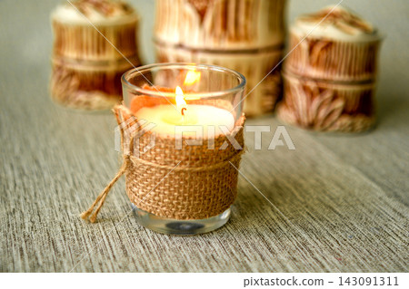 Rustic burlap-wrapped glass candle with warm flame on textured table for cozy home decor Rustic burlap-wrapped glass candle with warm flame on textured table for cozy home decor 143091311