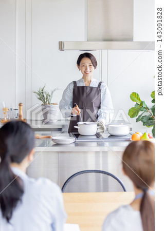 一位微笑的日本女教師在烹飪課上一邊攪拌鍋裡的食材一邊示範烹飪方法。 一位微笑的日本女教師在烹飪課上一邊攪拌鍋裡的食材一邊示範烹飪方法。 143091828