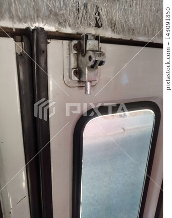 Close-up of a metal spring-loaded sliding door latch mounted with screws on a white door frame above a window with a black rubber seal and under decorative grey fringe. Close-up of a metal spring-loaded sliding door latch mounted with screws on a white door frame above a window with a black rubber seal and under decorative grey fringe. 143091850