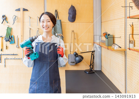 A smiling Japanese woman holding an electric drill and hammer in a DIY workshop. A smiling Japanese woman holding an electric drill and hammer in a DIY workshop. 143091913