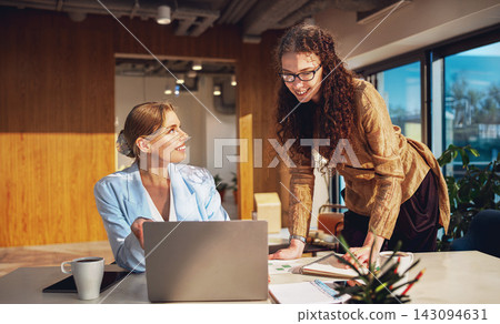 Enthusiastic mentor leans over the desk writing notes as her smiling blonde colleague looks up from a laptop Enthusiastic mentor leans over the desk writing notes as her smiling blonde colleague looks up from a laptop 143094631
