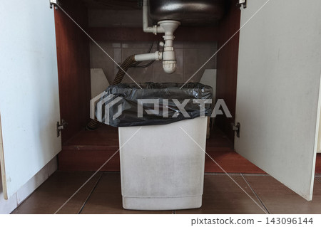 White household waste bin with a black plastic liner positioned beneath a kitchen sink, practical home waste disposal and cleaning setup. White household waste bin with a black plastic liner positioned beneath a kitchen sink, practical home waste disposal and cleaning setup. 143096144