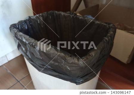 A large black liner neatly covers an open wastebasket indoors, household object used for everyday rubbish collection and domestic organization. A large black liner neatly covers an open wastebasket indoors, household object used for everyday rubbish collection and domestic organization. 143096147
