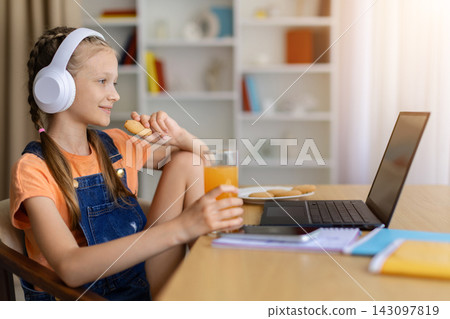 Cheerful preteen girl in headphones eating snacks while watching laptop at home 143097819