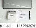 Top view of closed laptop, 2027 calendar showing January, pen and paper clip on white desk with sunlight shadows. Annual planning and event scheduling 143098979