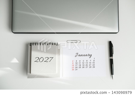 Top view of closed laptop, 2027 calendar showing January, pen and paper clip on white desk with sunlight shadows. Annual planning and event scheduling Top view of closed laptop, 2027 calendar showing January, pen and paper clip on white desk with sunlight shadows. Annual planning and event scheduling 143098979