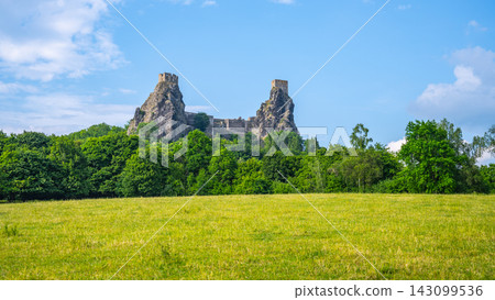 Trosky castle ruins rise above a green field in Bohemian Paradise. The structure is surrounded by trees and set against a blue sky. The scene shows nature and history together. 143099536