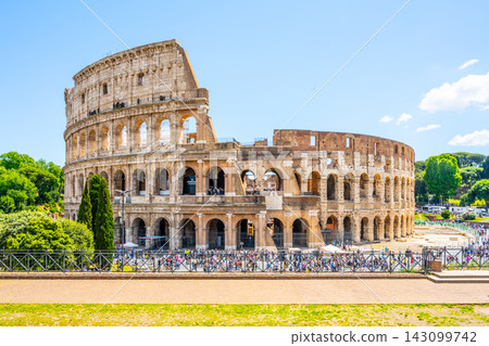 Tourists walk around the Colosseum in Rome. The ancient structure stands tall with its arches open to visitors. The scene shows clear skies and greenery in the background. Tourists walk around the Colosseum in Rome. The ancient structure stands tall with its arches open to visitors. The scene shows clear skies and greenery in the background. 143099742