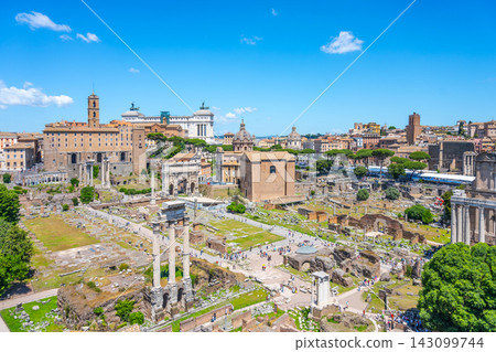 Tourists walk among ancient ruins at the Roman Forum in Rome Italy. The historic site shows remains of buildings and columns under a blue sky with few clouds. Tourists walk among ancient ruins at the Roman Forum in Rome Italy. The historic site shows remains of buildings and columns under a blue sky with few clouds. 143099744
