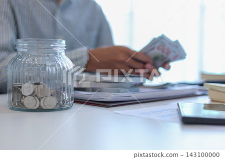 Close up glass jars filled with money and coins with businessman counting money in background. Close up glass jars filled with money and coins with businessman counting money in background. 143100000