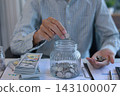 Businessman at working desk putting coins into a glass jar for saving money. Financial planning and saving money concept. 143100007