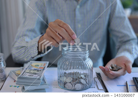 Businessman at working desk putting coins into a glass jar for saving money. Financial planning and saving money concept. Businessman at working desk putting coins into a glass jar for saving money. Financial planning and saving money concept. 143100007