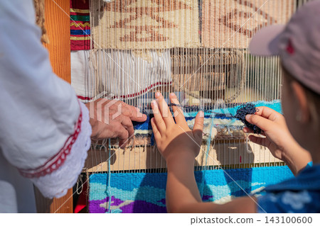 Child Learning Traditional Carpet Weaving at Loom in Kotel, Bulgaria. Child touches yarn while learning traditional carpet weaving from adult at wooden loom during outdoor cultural event Child Learning Traditional Carpet Weaving at Loom in Kotel, Bulgaria. Child touches yarn while learning traditional carpet weaving from adult at wooden loom during outdoor cultural event 143100600