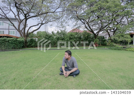 Asian man sits cross legged on a green lawn beneath large trees. He looks away while relaxing in a quiet garden. Asian man sits cross legged on a green lawn beneath large trees. He looks away while relaxing in a quiet garden. 143104039
