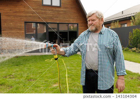 Senior man uses a garden hose to water the lawn outside a suburban home, illustrating active gardening, household maintenance, and outdoor lifestyle. Senior man uses a garden hose to water the lawn outside a suburban home, illustrating active gardening, household maintenance, and outdoor lifestyle. 143104304