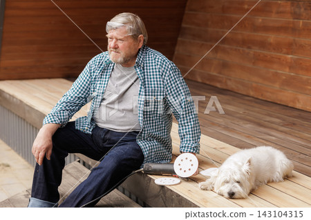 A senior homeowner pauses beside his sanding tool while a small dog rests on a wooden outdoor deck during terrace restoration. A senior homeowner pauses beside his sanding tool while a small dog rests on a wooden outdoor deck during terrace restoration. 143104315