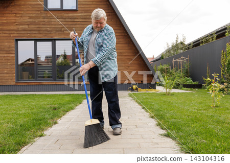 Elderly man sweeps a paved yard pathway beside his house, illustrating everyday home maintenance, outdoor cleaning, and senior household routines for lifestyle use. Elderly man sweeps a paved yard pathway beside his house, illustrating everyday home maintenance, outdoor cleaning, and senior household routines for lifestyle use. 143104316