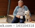 Senior man sits with his pet dog on a wooden porch, holding coffee during a quiet moment of companionship at home. 143104318