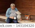Senior homeowner sits with his dog beside a paint bucket and brush on a wooden deck, illustrating outdoor terrace maintenance and refinishing. 143104320