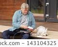Senior homeowner uses an orbital sander to refinish an outdoor wooden deck while his dog rests nearby, illustrating hands-on home maintenance and renovation. 143104325