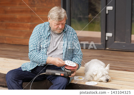 Senior homeowner uses an orbital sander to refinish an outdoor wooden deck while his dog rests nearby, illustrating hands-on home maintenance and renovation. Senior homeowner uses an orbital sander to refinish an outdoor wooden deck while his dog rests nearby, illustrating hands-on home maintenance and renovation. 143104325