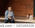 A mature homeowner sands an outdoor wooden deck with an orbital tool while his dog watches, illustrating DIY renovation, maintenance, and pet-friendly home improvement. 143104326