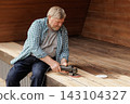 Older homeowner uses a power sander to restore weathered wooden decking during a terrace renovation project, illustrating hands-on maintenance and home improvement. 143104327