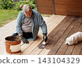 Senior homeowner applies protective oil to wooden deck boards while a dog rests nearby, illustrating terrace maintenance and outdoor home improvement. 143104329