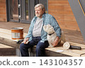 Senior homeowner sits with his dog on a wooden deck beside sanding equipment and coating supplies during an outdoor renovation project. 143104357