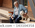 Senior homeowner holds a brush beside a dog during wooden deck restoration, illustrating outdoor maintenance, DIY refinishing, and pet companionship. 143104358