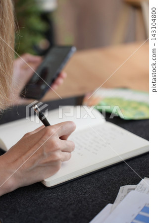 Woman writing expenses while checking smartphone at home Woman writing expenses while checking smartphone at home 143104800