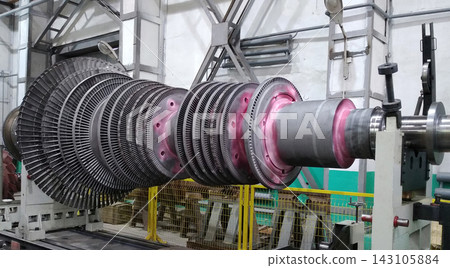 Large multi-stage steam turbine rotor with metal blades mounted on a heavy shaft inside a factory workshop, undergoing inspection and maintenance in a power plant manufacturing facility. Large multi-stage steam turbine rotor with metal blades mounted on a heavy shaft inside a factory workshop, undergoing inspection and maintenance in a power plant manufacturing facility. 143105884