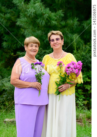Two smiling senior russian sisters with dental crowns holding flower bouquets standing together in a summer garden in Russia Two smiling senior russian sisters with dental crowns holding flower bouquets standing together in a summer garden in Russia 143106975
