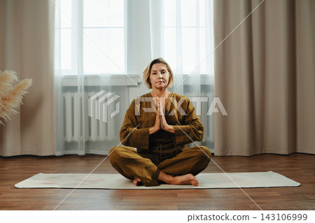 Middle-aged woman sits on yoga mat with hands in prayer position at home Middle-aged woman sits on yoga mat with hands in prayer position at home 143106999