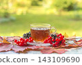 Transparent cup of tea and berries on a table covered by red maple leaves against green natural background; close up 143109690