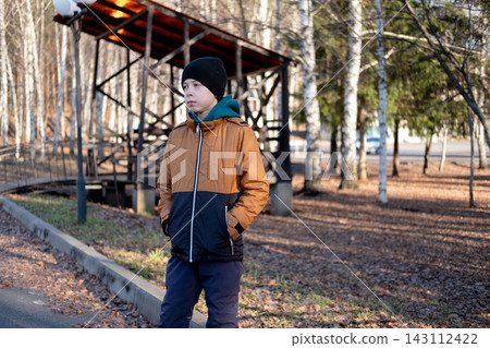 young caucasian boy with hands in pockets wearing a brown winter jacket standing on a park path near a wooden alcove or gazebo. young caucasian boy with hands in pockets wearing a brown winter jacket standing on a park path near a wooden alcove or gazebo. 143112422
