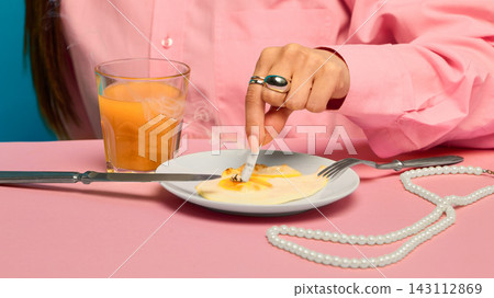 Woman in pink shirt stubbing cigarette into fried egg beside juice glass and pearl necklace. 143112869