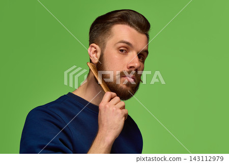 Bearded man holding wooden comb near face on green background. 143112979