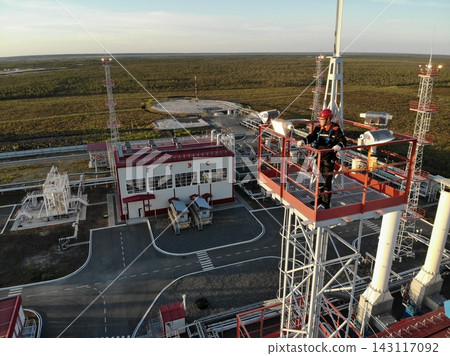 Industrial worker wearing hard hat and harness stands among elevated processing equipment at remote facility. Tall lattice towers, pipes, access platforms, and flat northern terrain extend behind him. Industrial worker wearing hard hat and harness stands among elevated processing equipment at remote facility. Tall lattice towers, pipes, access platforms, and flat northern terrain extend behind him. 143117092