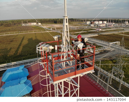 Industrial worker in safety helmet checks mounted lights on elevated tower platform, focused and alert. Remote refinery style site spreads below with pipes, access roads, and forest edge. Industrial worker in safety helmet checks mounted lights on elevated tower platform, focused and alert. Remote refinery style site spreads below with pipes, access roads, and forest edge. 143117094