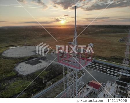 Communications tower rises above industrial compound as mounted signal equipment faces surrounding terrain. Sunset light illuminates fenced platforms, service roads, utility structures Communications tower rises above industrial compound as mounted signal equipment faces surrounding terrain. Sunset light illuminates fenced platforms, service roads, utility structures 143117096
