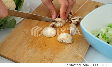 Chef practices knife safety while slicing button mushroom on bamboo board in bright kitchen, demonstrating safe finger positioning alongside fresh cauliflower, celery, and salad greens. Chef practices knife safety while slicing button mushroom on bamboo board in bright kitchen, demonstrating safe finger positioning alongside fresh cauliflower, celery, and salad greens. 143117113