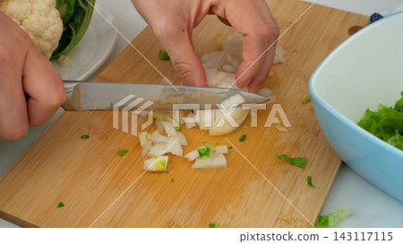 Cook slices fresh white onion using kitchen knife on bamboo cutting board during culinary food prep. Focused hands chop fresh vegetables carefully beside bowl of salad greens and cauliflower. Cook slices fresh white onion using kitchen knife on bamboo cutting board during culinary food prep. Focused hands chop fresh vegetables carefully beside bowl of salad greens and cauliflower. 143117115